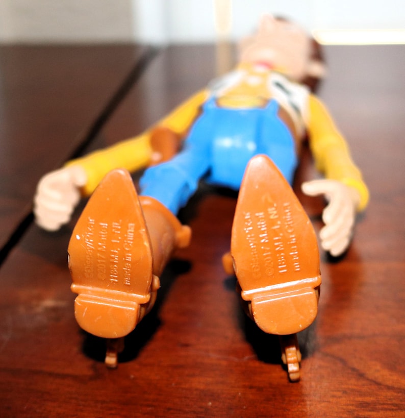 Puede incluir: Figura de juguete de un vaquero tumbado boca arriba. La figura lleva una camisa amarilla, un peto azul y botas marrones. Se ven las suelas de las botas, con el texto "&copy;2017 Mattel 1186 MJ, 1, NC, made in China."