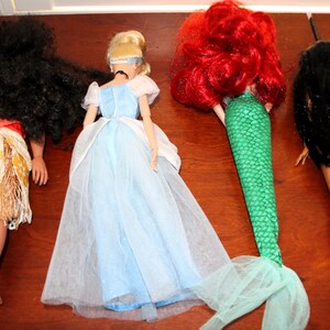 May include: Four dolls viewed from behind. The first has long black hair and a tan outfit with a floral pattern. The second has blonde hair and a light blue dress. The third has bright red hair and a green mermaid tail. The fourth has black hair.