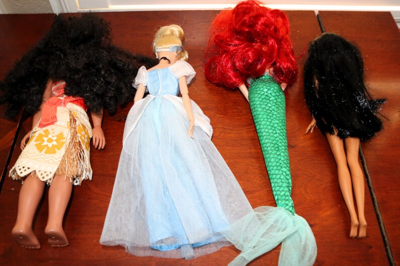 May include: Four dolls viewed from behind. The first has long black hair and a tan outfit with a floral pattern. The second has blonde hair and a light blue dress. The third has bright red hair and a green mermaid tail. The fourth has black hair.