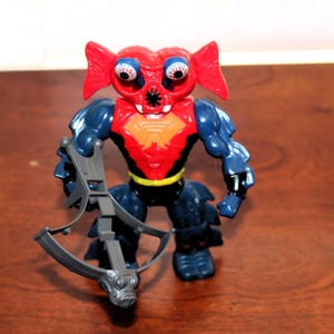 May include: A plastic action figure with a red head, large blue eyes with red veins, and prominent red ear-like fins. The torso is red with an orange chest plate, while the arms and legs are dark blue. It holds a gray plastic crossbow accessory.