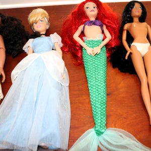 May include: Four fashion dolls arranged in a row. The first doll has dark curly hair and a patterned dress. The second doll has blonde hair and a light blue gown. The third doll has red hair and a green mermaid tail. The fourth doll has long black hair.
