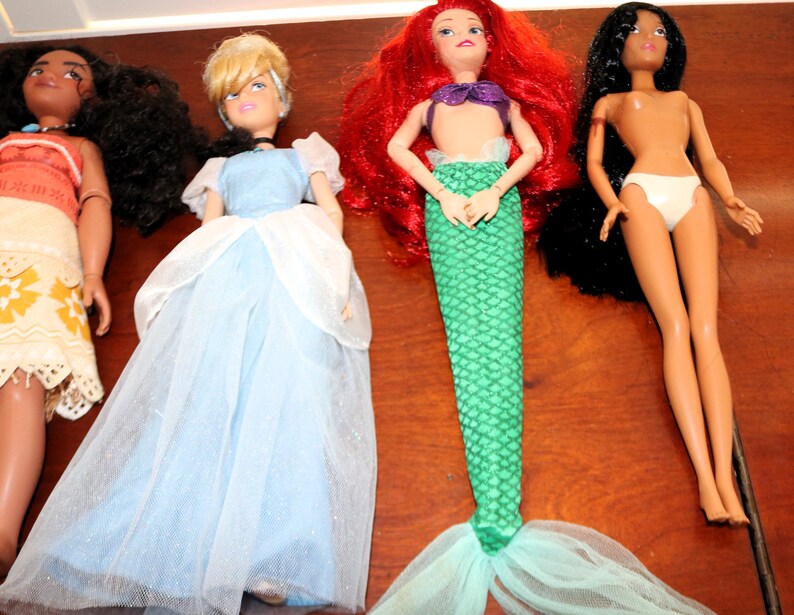 May include: Four fashion dolls arranged in a row. The first doll has dark curly hair and a patterned dress. The second doll has blonde hair and a light blue gown. The third doll has red hair and a green mermaid tail. The fourth doll has long black hair.