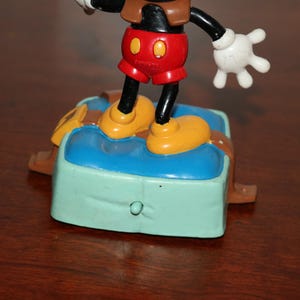 May include: A Mickey Mouse figurine standing on a blue and green base with brown accents. Mickey wears red shorts, yellow shoes, and white gloves. The figurine is a collectible item.