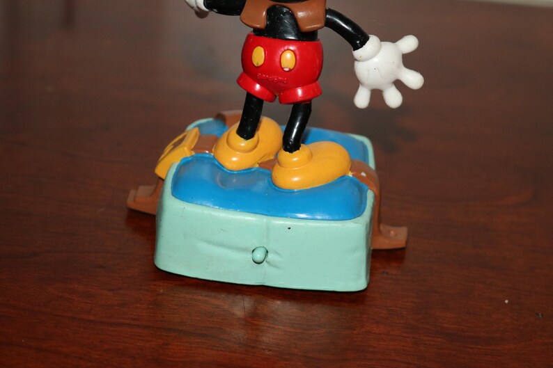May include: A Mickey Mouse figurine standing on a blue and green base with brown accents. Mickey wears red shorts, yellow shoes, and white gloves. The figurine is a collectible item.