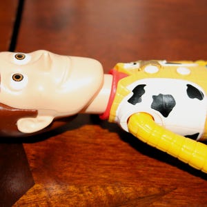 Puede incluir: Una figura de acci&oacute;n de Woody, con una cara bronceada y cabello casta&ntilde;o, est&aacute; sobre una superficie de madera. La figura lleva un chaleco amarillo con un estampado de piel de vaca en blanco y negro, un cuello rojo y una camisa azul. El brazo del juguete es amarillo.