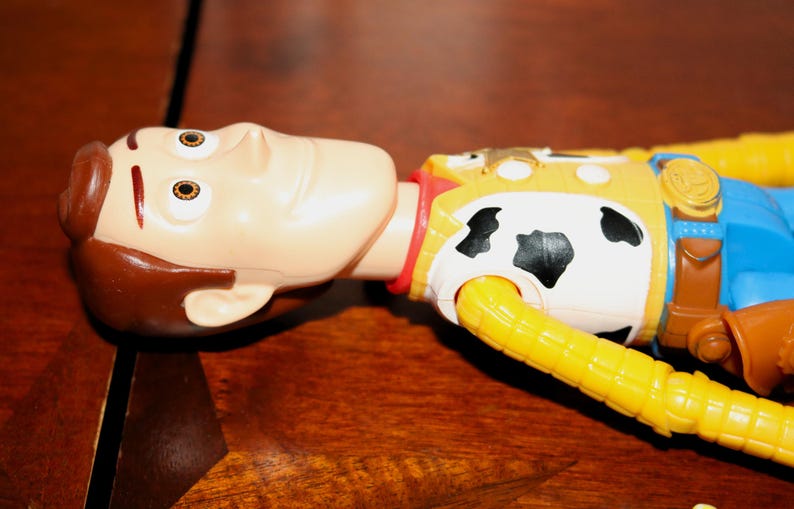 Puede incluir: Una figura de acci&oacute;n de Woody, con una cara bronceada y cabello casta&ntilde;o, est&aacute; sobre una superficie de madera. La figura lleva un chaleco amarillo con un estampado de piel de vaca en blanco y negro, un cuello rojo y una camisa azul. El brazo del juguete es amarillo.