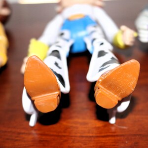 Puede incluir: Primer plano de las botas marrones de un personaje de juguete con texto visible. La figura tiene una camisa blanca, un chaleco azul y pantalones con estampado en blanco y negro. Las botas son de color marr&oacute;n claro.
