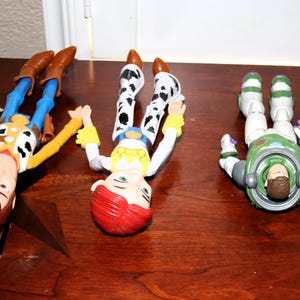 Puede incluir: Tres figuras de personajes de Toy Story: Woody, Jessie y Buzz Lightyear, junto con una peque&ntilde;a figura de gato amarillo, est&aacute;n dispuestas sobre una superficie de madera marr&oacute;n. Woody viste un atuendo de vaquero, Jessie tiene un look de vaquera y Buzz lleva un traje espacial.