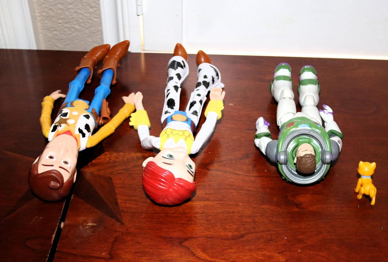 Puede incluir: Tres figuras de personajes de Toy Story: Woody, Jessie y Buzz Lightyear, junto con una peque&ntilde;a figura de gato amarillo, est&aacute;n dispuestas sobre una superficie de madera marr&oacute;n. Woody viste un atuendo de vaquero, Jessie tiene un look de vaquera y Buzz lleva un traje espacial.