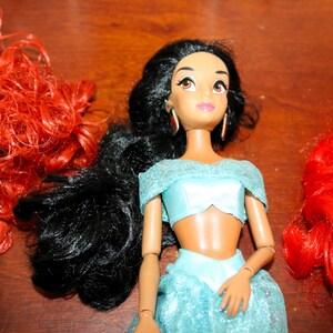 Puede incluir: Mu&ntilde;eca de la princesa Jasmine con pelo largo y negro, vestida con un atuendo azul claro y pendientes dorados. Se ve pelo rojo a ambos lados de la mu&ntilde;eca. La mu&ntilde;eca tiene ojos marrones y piel clara.