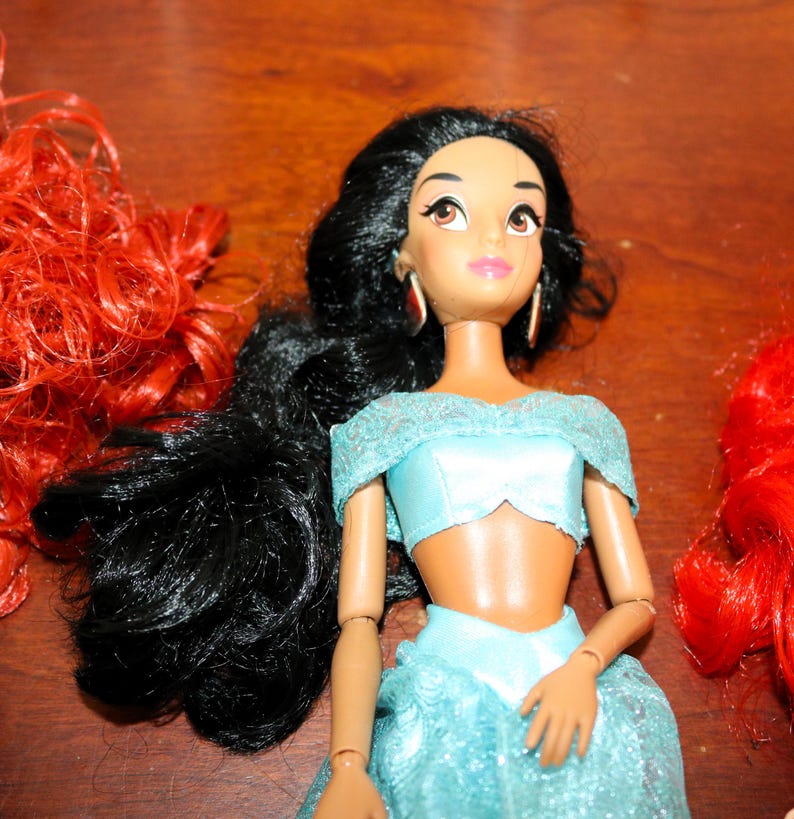 Puede incluir: Mu&ntilde;eca de la princesa Jasmine con pelo largo y negro, vestida con un atuendo azul claro y pendientes dorados. Se ve pelo rojo a ambos lados de la mu&ntilde;eca. La mu&ntilde;eca tiene ojos marrones y piel clara.