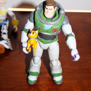 Puede incluir: Figura de acci&oacute;n de Buzz Lightyear con un traje espacial verde y blanco, sosteniendo una peque&ntilde;a figura de gato amarillo. La figura tiene una cabeza marr&oacute;n y un casco verde. El gato tiene un collar azul.