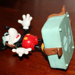 May include: A Mickey Mouse figurine in a lying down position. The figure has a black body, white gloves, and a red shorts. A light green box is attached to the figure with brown straps.
