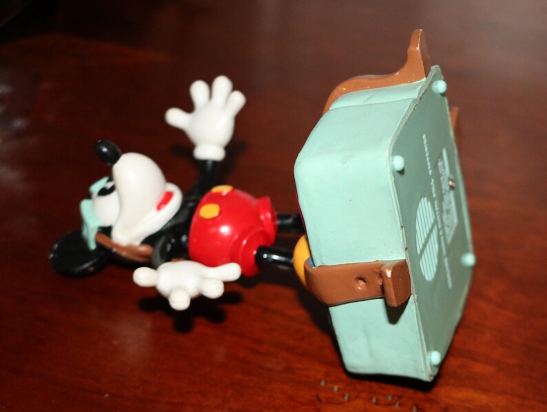 May include: A Mickey Mouse figurine in a lying down position. The figure has a black body, white gloves, and a red shorts. A light green box is attached to the figure with brown straps.