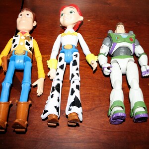 Puede incluir: Tres figuras de acci&oacute;n de personajes de Toy Story. Woody con camisa amarilla, pantalones azules y botas marrones. Jessie con top blanco, cintur&oacute;n azul y pantalones con estampado de vaca. Buzz Lightyear con traje espacial blanco y verde, y un peque&ntilde;o gato amarillo.