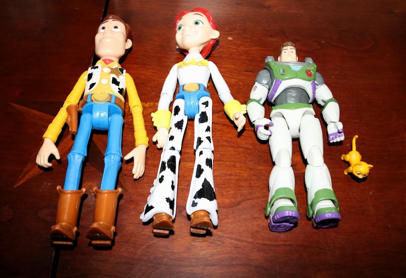 Puede incluir: Tres figuras de acci&oacute;n de personajes de Toy Story. Woody con camisa amarilla, pantalones azules y botas marrones. Jessie con top blanco, cintur&oacute;n azul y pantalones con estampado de vaca. Buzz Lightyear con traje espacial blanco y verde, y un peque&ntilde;o gato amarillo.