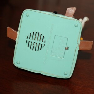May include: A light blue, square-shaped toy base with a speaker grill, battery compartment, and brown tabs. The base has a few screws and is made of plastic. The text on the base is blurry.