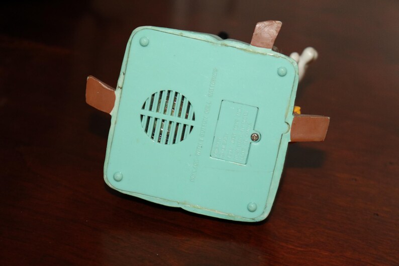 May include: A light blue, square-shaped toy base with a speaker grill, battery compartment, and brown tabs. The base has a few screws and is made of plastic. The text on the base is blurry.