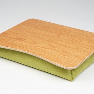 Puede incluir: Un escritorio de regazo de madera con un cojín de tela verde. El escritorio está curvado para adaptarse al regazo y tiene un acabado de madera natural.