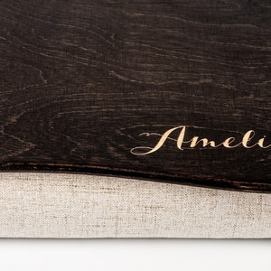 Op de afbeelding: Een donkerbruine houten opbergdoos met een voering van linnen stof. De doos heeft een gebogen rand en de naam "Amelia" is in gouden letters gegraveerd op de bovenkant.