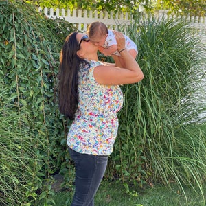 May include: A woman wearing sunglasses and a colorful crochet top is holding a baby in her arms and kissing the baby on the cheek. The baby is wearing a white short-sleeved shirt with a colorful pattern.