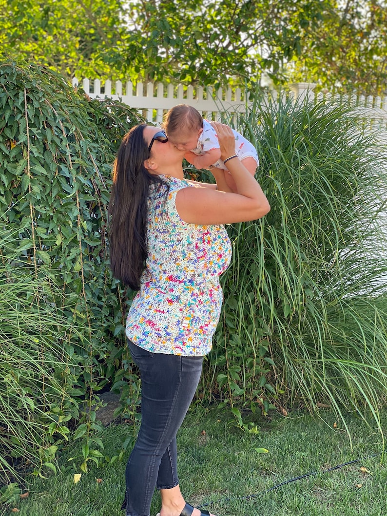 May include: A woman wearing sunglasses and a colorful crochet top is holding a baby in her arms and kissing the baby on the cheek. The baby is wearing a white short-sleeved shirt with a colorful pattern.