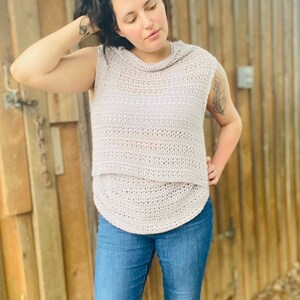 May include: A light gray crochet top with a cowl neck and sleeveless design. The top is made with a delicate open stitch pattern and has a relaxed fit.