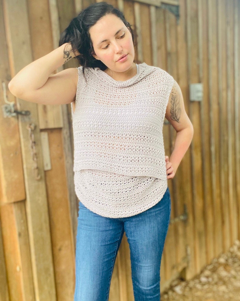May include: A light gray crochet top with a cowl neck and sleeveless design. The top is made with a delicate open stitch pattern and has a relaxed fit.