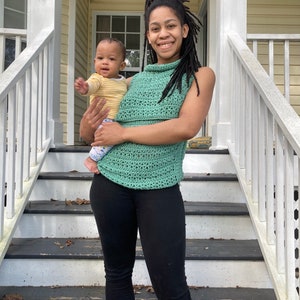 May include: A woman wearing a green crochet top and black pants is holding a baby wearing a yellow and white striped shirt. The woman and baby are standing on a set of wooden stairs with a white railing.