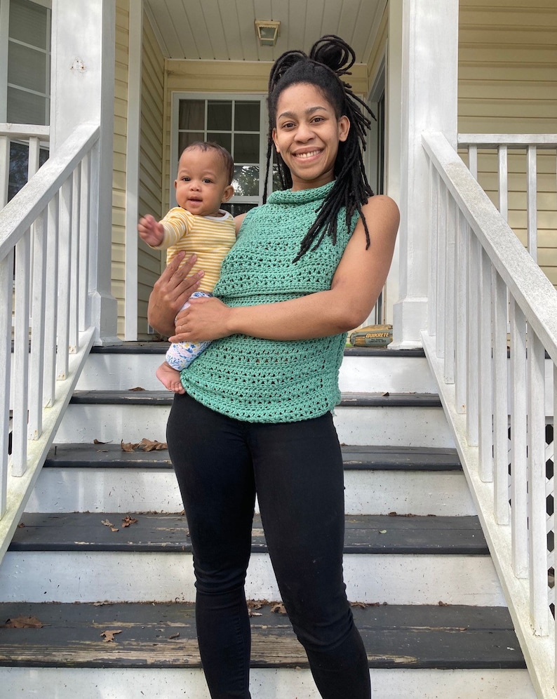 May include: A woman wearing a green crochet top and black pants is holding a baby wearing a yellow and white striped shirt. The woman and baby are standing on a set of wooden stairs with a white railing.