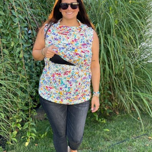 May include: A woman wearing a colorful floral print crochet top with a black tank top underneath, black jeans, and black sandals. She is standing in front of a green leafy background.