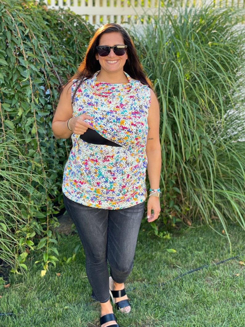 May include: A woman wearing a colorful floral print crochet top with a black tank top underneath, black jeans, and black sandals. She is standing in front of a green leafy background.
