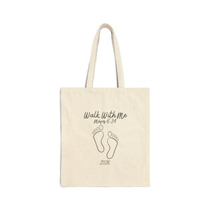 May include: Cream-colored tote bag with black text and a graphic of footprints. The text reads "Walk With Me Moses 6:34" above the footprints and "2026" below. The bag has two handles.