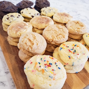 May include: A wooden serving board displays an assortment of freshly baked cookies. Varieties include chocolate chip, cinnamon sugar, and sprinkle-covered cookies. The cookies are arranged in stacks and rows, showcasing their textures and colors.