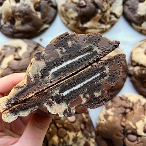 May include: A close-up of a chocolate cookie cut in half, revealing a white cream filling. The cookie is studded with chocolate chips and has a marbled appearance. Several other cookies are visible in the background.