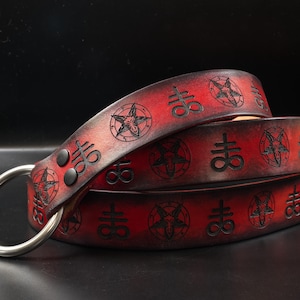 May include: A red and black gradient leather belt featuring embossed black pentagram and Leviathan cross symbols. The belt is secured with two black rivets and includes a large, circular silver-toned metal ring at one end for fastening.