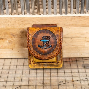 May include: A brown leather case with a stitched design and an anchor with a blue rope motif. The case is on a gridded surface.