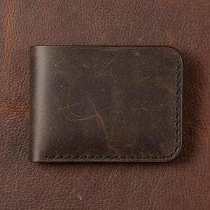 May include: A dark brown leather wallet with a slightly distressed finish. The wallet is rectangular with rounded corners and features black stitching along the edges. The leather has a textured appearance.