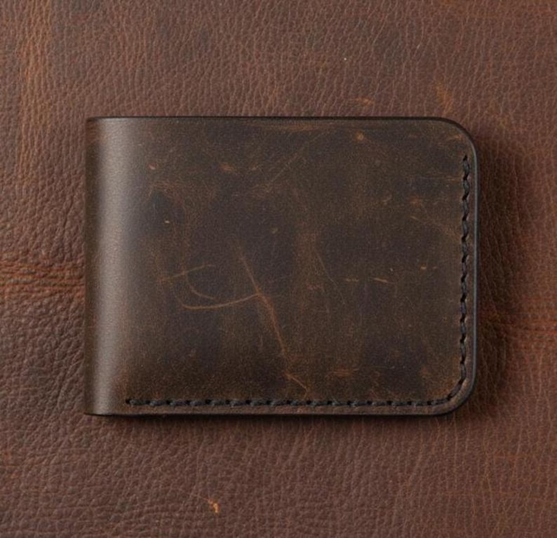 May include: A dark brown leather wallet with a slightly distressed finish. The wallet is rectangular with rounded corners and features black stitching along the edges. The leather has a textured appearance.