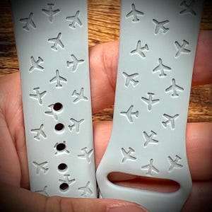 May include: Two light gray watch bands with a pattern of small airplanes. The bands are held in a person's hand. The bands have several holes for adjustment and a buckle. The watch bands are made of a flexible material.