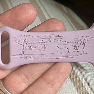 May include: A light purple silicone watch band with an engraved design of a savanna scene with trees, a moon, and two hippos.
