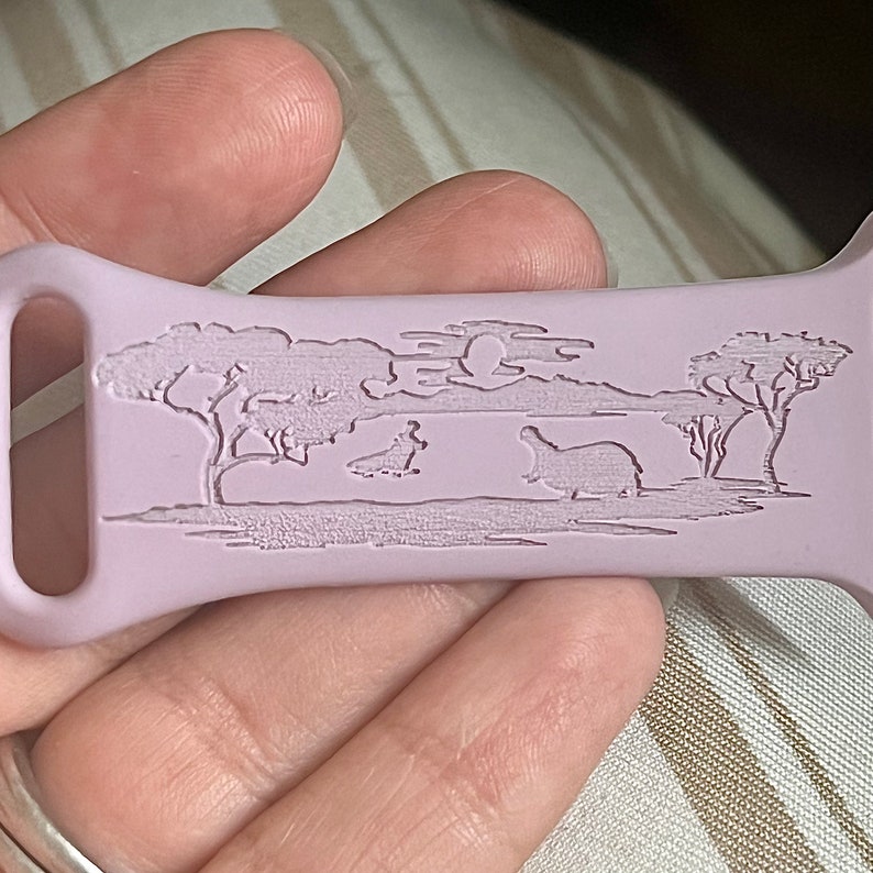 May include: A light purple silicone watch band with an engraved design of a savanna scene with trees, a moon, and two hippos.