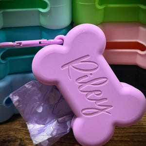 May include: A light purple, bone-shaped silicone waste bag dispenser featuring the name Riley embossed in a script font on the front. The dispenser includes a matching light purple carabiner clip and a small portion of a light purple patterned bag.