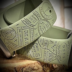 May include: A green Apple Watch band with a travel theme. The band is decorated with embossed stamps from various countries, including Paris, France, Brazil, Jamaica, and Australia. The stamps feature dates, arrival and departure information, and other travel-related details.