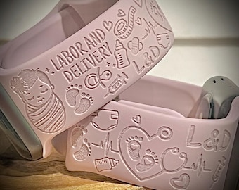 May include: Two pink silicone Apple Watch bands with engraved designs. The top band has the text "Labor and Delivery" with a baby bottle, a pacifier, a syringe, and footprints. The bottom band has a heart with footprints, a stethoscope, and the text "L&D".