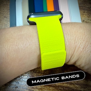 May include: A bright neon yellow magnetic watch band is displayed on a wrist. The band is attached to a silver watch face. The text "MAGNETIC BANDS" is in a white font on a black background. Other colored bands are visible in the background.