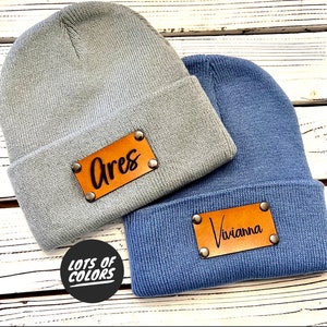 May include: Two knitted beanies with leather patches. The beanie on the left is light blue with a brown leather patch that says "Ares". The beanie on the right is blue with a brown leather patch that says "Viviana".