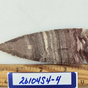 May include: A stone arrowhead with a layered pattern of purple, gray, and white. The arrowhead is placed next to a wooden ruler with inch measurements. A blue and white label below the arrowhead reads "26104S4-4".