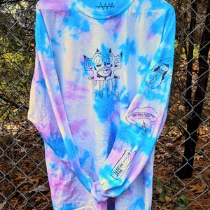 May include: A long-sleeved tie-dye shirt in shades of blue, pink, and white with black ink illustrations of a hand with an eye, a mouth, a phone, and a bag of money with flames. The shirt has the text "AWW" printed on the neck.