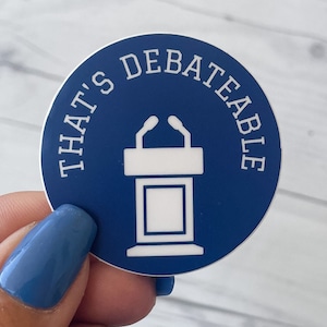 Op de afbeelding: Een ronde sticker met een donkerblauwe achtergrond en een witte afbeelding van een spreekgestoelte met twee microfoons. De tekst "THAT'S DEBATEABLE" is in witte hoofdletters in een boog langs de bovenrand van het stickerontwerp gedrukt.